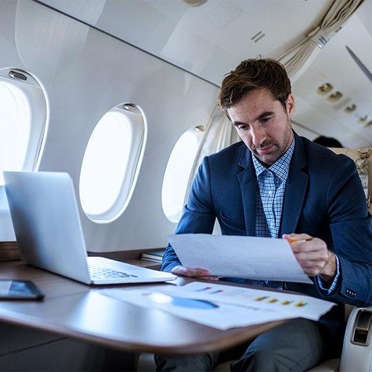 Man sitting at a table in a private jet working on his laptop and reading paperwork