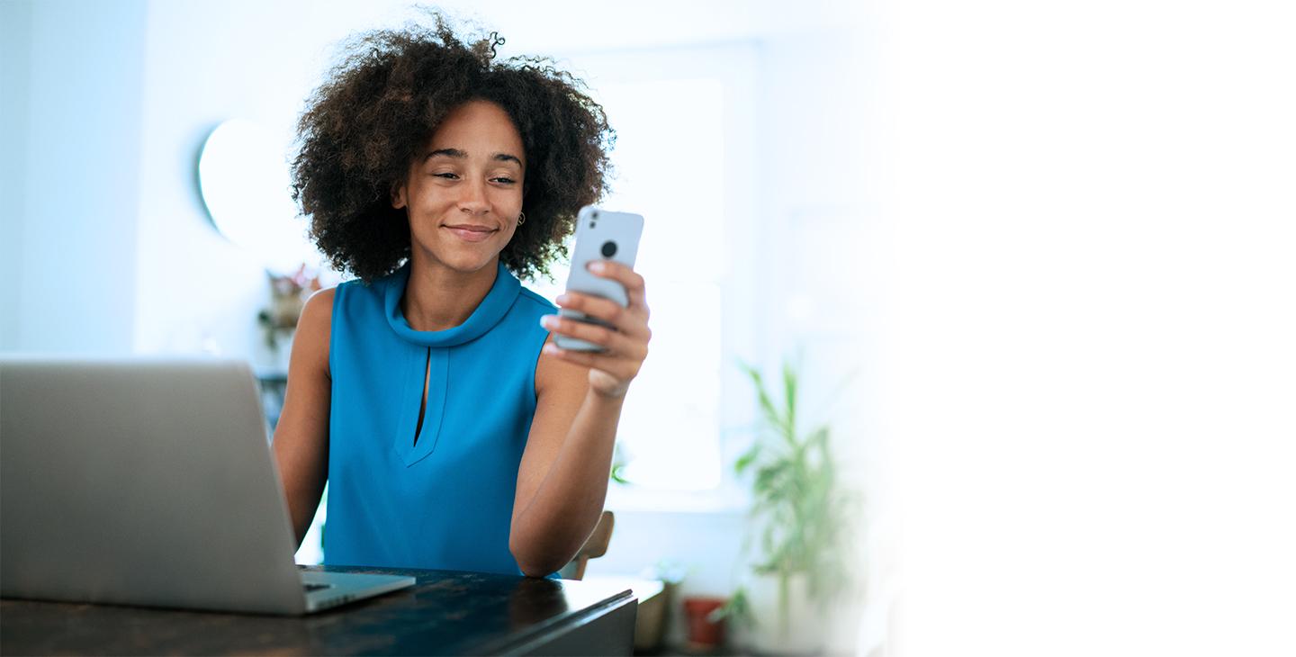 穿蓝衬衫的女人, 在笔记本电脑前微笑着看她的手机, 享受Viasat的互联网套餐